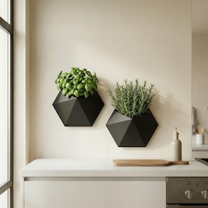 May include: Two black, geometric wall planters, each filled with fresh herbs, are mounted on a light-colored wall. One planter holds basil, the other rosemary. The planters are above a white countertop in a kitchen setting.