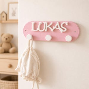 May include: A pink oval wall-mounted coat rack featuring the white raised name LUKAS in a decorative font. The rack includes three white cylindrical hooks, with one hook holding a cream-coloured fabric bag with a drawstring closure.