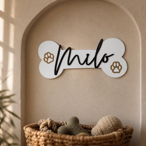 Può includere: Una targa da parete bianca a forma di osso con il nome Milo in caratteri corsivi neri. Due ritagli di impronte di zampe color oro sono posizionati sui lati sinistro e destro della forma a osso, creando una decorazione da parete a tema animali.
