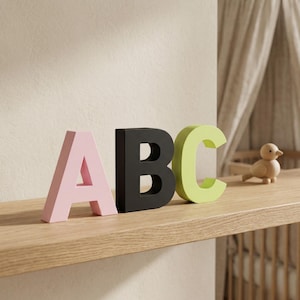Op de afbeelding: Houten letters 'ABC' in roze, zwart en groen, geplaatst op een lichte houten plank. Een klein houten vogelbeeldje staat ernaast. De letters zijn een decoratief element, mogelijk voor een kinderkamer.