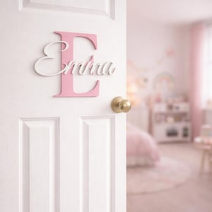 May include: A pink wooden letter "E" with the name "Emma" in white script is mounted on a white door. The door has a gold doorknob. The background shows a blurred pink bedroom.