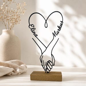 Può includere: Una scultura in metallo nero che raffigura due mani che si tengono, formando una forma a cuore nella parte superiore. Il nome Elina è integrato sul lato sinistro del cuore e il nome Markus sul lato destro. Poggia su una base rettangolare in legno.
