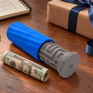 May include: A cylindrical gray puzzle box featuring a complex maze pattern on its exterior, partially encased in a bright blue silicone sleeve. A rolled twenty-dollar bill is positioned next to the puzzle container on a wooden surface.