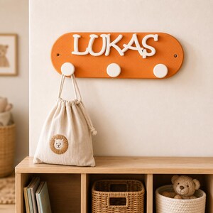Op de afbeelding: Een oranje ovale wandkapstok met drie witte ronde haken en de witte naam LUKAS aan de voorzijde bevestigd. Een beige stoffen tas met trekkoord en een kleine geborduurde leeuwengezicht-afbeelding hangt aan een van de witte haken.