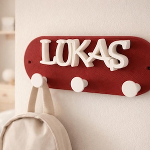 May include: A red wall-mounted coat rack with the name "LUKAS" in white, three white hooks, and a beige backpack hanging from one hook. The rack is mounted on a white wall.