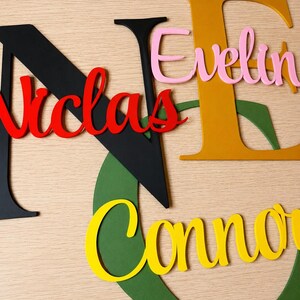 May include: A collection of layered wooden letters and names in various colors. Large black letter N with red script text Nicolas, large mustard yellow letter E with pink script text Evelyn, and a large green letter G with yellow script text Connor.