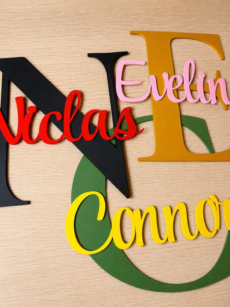 May include: A collection of layered wooden letters and names in various colors. Large black letter N with red script text Nicolas, large mustard yellow letter E with pink script text Evelyn, and a large green letter G with yellow script text Connor.
