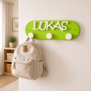 Puede incluir: Un perchero de pared ovalado de color verde lima con el nombre LUKAS en relieve blanco y tres ganchos circulares blancos. Una mochila de lona beige con un bolsillo frontal con cremallera y detalles de piel sintética marrón cuelga de un gancho.