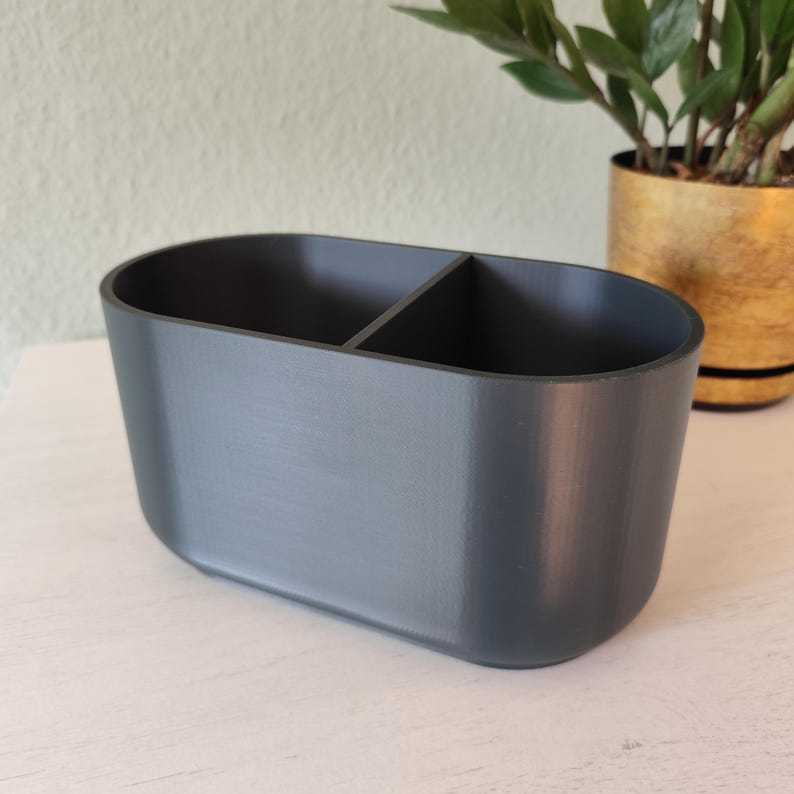 May include: A gray, oval-shaped organizer with two compartments. The container is made of a solid material and has rounded edges. The background includes a plant in a gold pot.