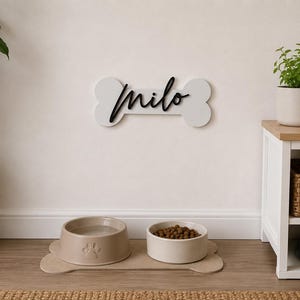 Può includere: Una decorazione da parete a forma di osso bianca con il nome "Milo" in corsivo nero. Sotto, due ciotole per cani sono su un tappetino a forma di osso. Una foto incorniciata di un golden retriever e una pianta in vaso sono su un tavolino.