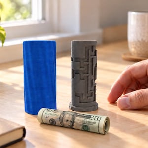 Könnte beinhalten: Ein zylindrischer blauer Behälter und eine graue Puzzle-Box mit Labyrinth-Muster auf der Außenseite, begleitet von einem gerollten Zwanzig-Dollar-Schein. Die graue Puzzle-Box hat einen runden Boden und einen komplexen Pfad auf der Oberfläche.