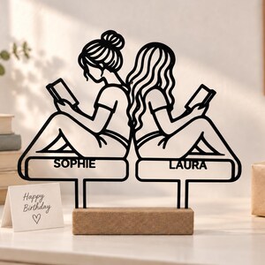 Può includere: Una sagoma in metallo nero di due persone sedute schiena contro schiena mentre leggono libri, montata su una base rettangolare in legno. I nomi SOPHIE e LAURA sono incisi sui sedili. Un piccolo biglietto riporta la scritta Happy Birthday.