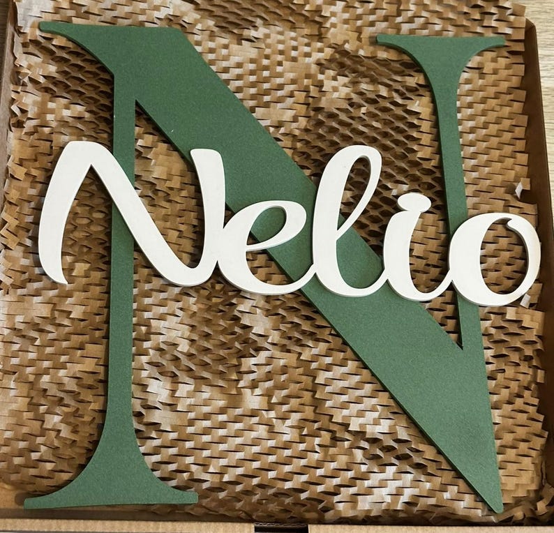 May include: A large, matte sage green wooden letter N with a white script-style wooden cutout of the name Nelio layered on top. The letters feature smooth edges and a flat finish, creating a layered, dimensional appearance for the custom sign.