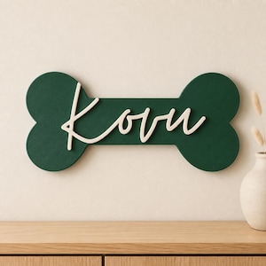 Könnte beinhalten: Ein dunkelgrünes, knochenförmiges Wandschild mit dem Namen Kovu in erhabener, weißer Schreibschrift. Das Schild hat eine texturierte Oberfläche und ist horizontal montiert, wobei der Name deutlich in der Mitte der Knochenform zu sehen ist.