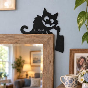 Puede incluir: Una decoración de pared de metal negro con forma de gato de dibujos animados con ojos grandes y una amplia sonrisa, sosteniendo un cuchillo de carnicero. El nombre LUNA está recortado en el cuerpo del gato. La pieza está sobre la esquina superior de un marco de espejo de madera.