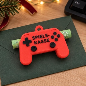 Könnte beinhalten: Ein leuchtend roter Geldhalter in Form eines Gamecontrollers mit einer kleinen Öse an der Oberseite. Der Controller hat schwarze Tasten und ein Steuerkreuz, mit dem schwarzen Schriftzug "SPIELE-KASSE" auf der Vorderseite. Ein gerollter Geldschein steckt in der Mitte.
