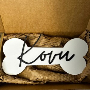 Personalisiertes Hunde-Namensschild Kovu auf weißem Knochen mit schwarzem 3D-Schriftzug, verpackt in einer Geschenkbox.