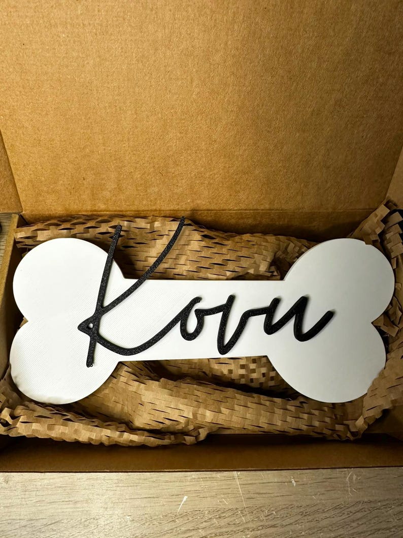 Personalisiertes Hunde-Namensschild Kovu auf weißem Knochen mit schwarzem 3D-Schriftzug, verpackt in einer Geschenkbox.