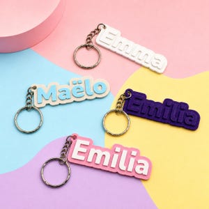 Könnte beinhalten: Vier rechteckige Schlüsselanhänger mit Metallringen. Einer ist weiß mit dem Namen Emma, einer beige mit hellblauem Text Maëlo, einer dunkelviolett mit dem Namen Emilia und einer hellrosa mit weißem Text Emilia in erhabener Schrift.