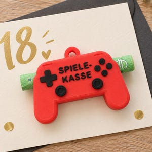 Könnte beinhalten: Eine cremefarbene Grußkarte mit der goldfarbenen Zahl 18 und einem kleinen Herz. Auf der Karte befindet sich ein roter Kunststoff-Controller als Anhänger mit schwarzen Tasten und dem schwarzen Schriftzug SPIELE-KASSE, der einen grünen Geldschein hält.