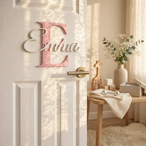 Op de afbeelding: Een roze houten letter "E" met de naam "Emma" in witte letters is op een witte deur gemonteerd. De deur heeft een goudkleurige handgreep. De scène is zacht verlicht, wat wijst op een kinderkamer.