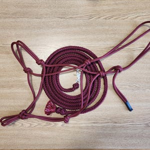 Op de afbeelding: Een bordeauxrood paardenhalster en touwset. Het halster is gemaakt van geweven touw en heeft een geknoopt ontwerp. Het touw is opgerold en bevestigd aan het halster met een zilverkleurige sluiting. De set wordt gepresenteerd op een lichtbruin houten oppervlak.