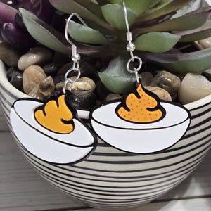 May include: A pair of earrings shaped like a white bowl with a yellow yolk inside. The earrings are made of white and yellow plastic and have silver hooks.
