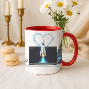 Puede incluir: Taza de cerámica blanca con interior y asa rojos. La taza presenta un diseño de un cohete que se lanza desde la pantalla de un portátil, con un código binario en forma de corazón en la parte superior. Un gráfico de placa de circuito está en el lateral.