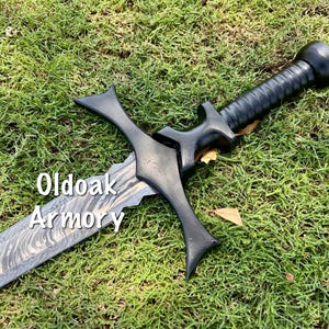 May include: A black sword with a patterned blade and a black handle lies on green grass. The sword has a cross-shaped guard and the text "Oldoak Armory" is visible on the blade.