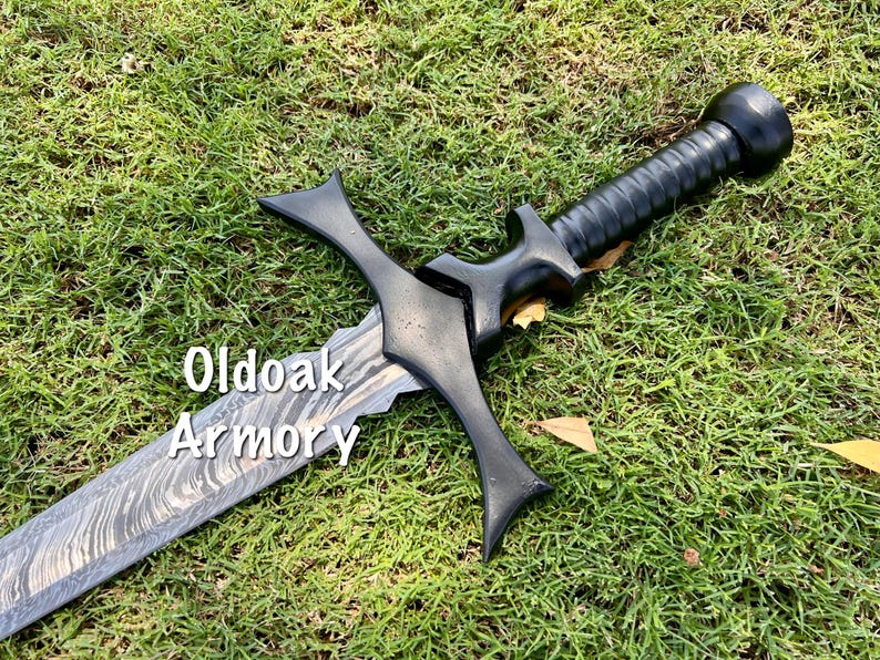 May include: A black sword with a patterned blade and a black handle lies on green grass. The sword has a cross-shaped guard and the text "Oldoak Armory" is visible on the blade.