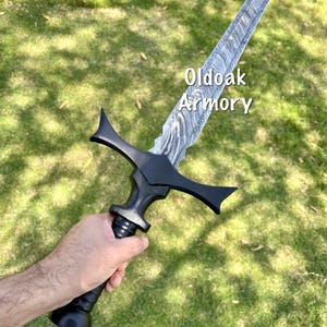 May include: A black sword with a patterned blade is held against a green grassy background. The sword has a black handle and crossguard. The text "Oldoak Armory" is visible on the blade.