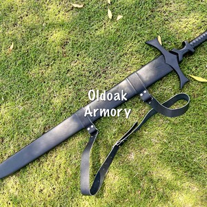 May include: A black sword with a cross-shaped hilt and a leather sheath, lying on green grass. The sword has a textured handle and a strap. The text "Oldoak Armory" is visible on the sheath.