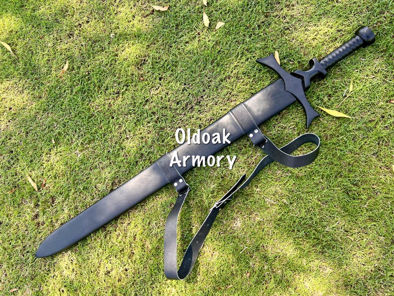 May include: A black sword with a cross-shaped hilt and a leather sheath, lying on green grass. The sword has a textured handle and a strap. The text "Oldoak Armory" is visible on the sheath.