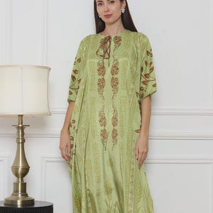 May include: A long, flowing, pale green dress with a brown floral pattern. The dress has short sleeves and a tie at the neckline. The hemline and sleeves have a brown border. The dress is worn with silver sandals.