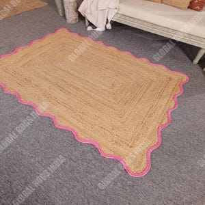 May include: A rectangular jute rug with a scalloped pink border. The rug has a natural tan color and a woven texture. The pink border adds a pop of color and a decorative element to the rug. This rug is suitable for living rooms and bedrooms.