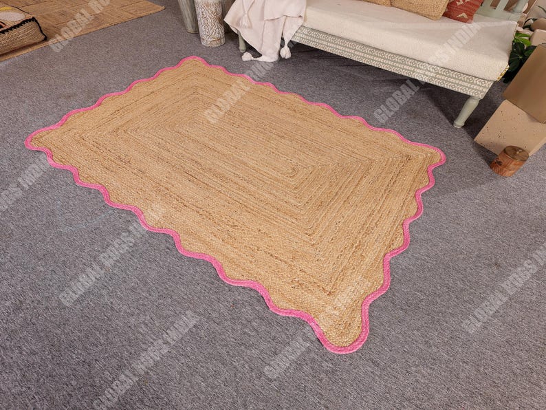 May include: A rectangular jute rug with a scalloped pink border. The rug has a natural tan color and a woven texture. The pink border adds a pop of color and a decorative element to the rug. This rug is suitable for living rooms and bedrooms.