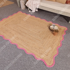 May include: A rectangular jute rug with a scalloped pink border. The rug has a natural tan color and a woven texture. The rug is placed on a gray surface. A small tray with cups and a wooden block are on the rug.