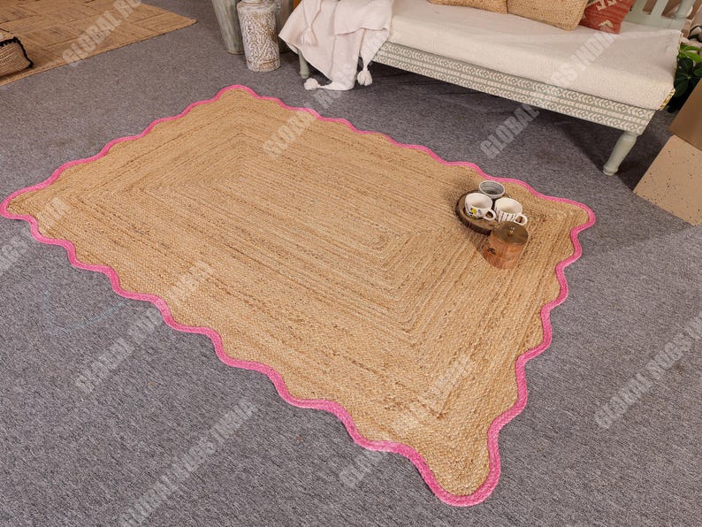 May include: A rectangular jute rug with a scalloped pink border. The rug has a natural tan color and a woven texture. The rug is placed on a gray surface. A small tray with cups and a wooden block are on the rug.