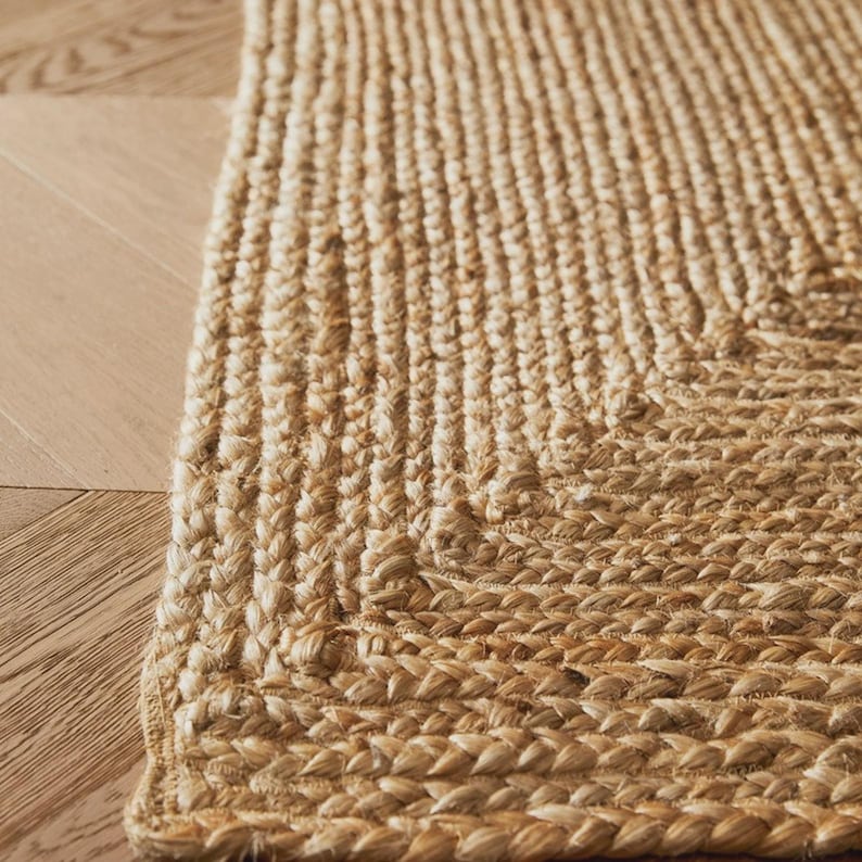 May include: Close-up of a natural jute rug with a braided border. The rug has a textured surface and a warm, neutral color, suitable for various interior styles. The rug is placed on a light-colored wooden floor.