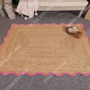May include: A rectangular jute rug with a scalloped pink border. The rug has a natural tan color and a woven texture. The pink border adds a pop of color and a decorative touch. The rug is suitable for indoor use.