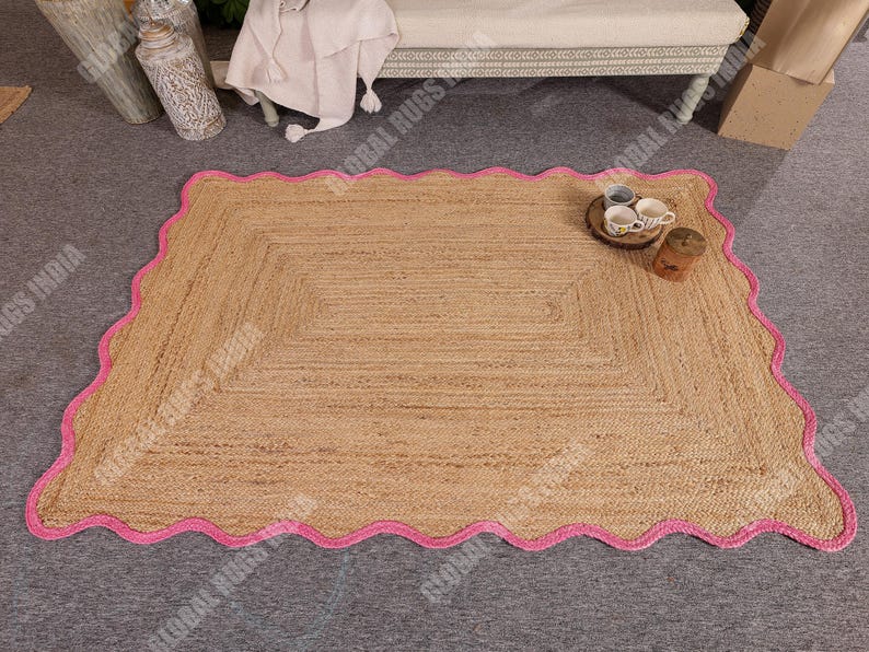 May include: A rectangular jute rug with a scalloped pink border. The rug has a natural tan color and a woven texture. The pink border adds a pop of color and a decorative touch. The rug is suitable for indoor use.