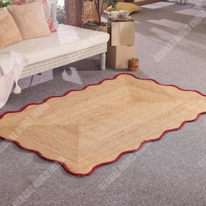 May include: A rectangular jute rug with a scalloped edge and a red border. The rug is light brown and sits on a gray carpet. The rug is a decorative floor covering.