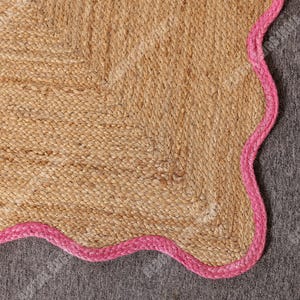 May include: A natural jute rug with a scalloped edge in a vibrant pink color. The rug has a woven texture and a neutral beige color, creating a contrast with the bright pink border. This rug is suitable for home decor.
