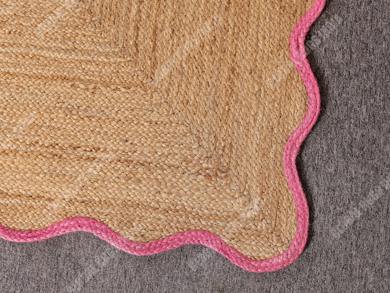 May include: A natural jute rug with a scalloped edge in a vibrant pink color. The rug has a woven texture and a neutral beige color, creating a contrast with the bright pink border. This rug is suitable for home decor.