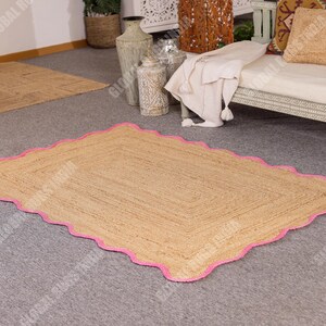 May include: A rectangular jute rug with a scalloped pink border. The rug has a natural tan color and is placed on a gray carpet. The rug's design features a woven texture, adding a rustic touch to the room's decor.