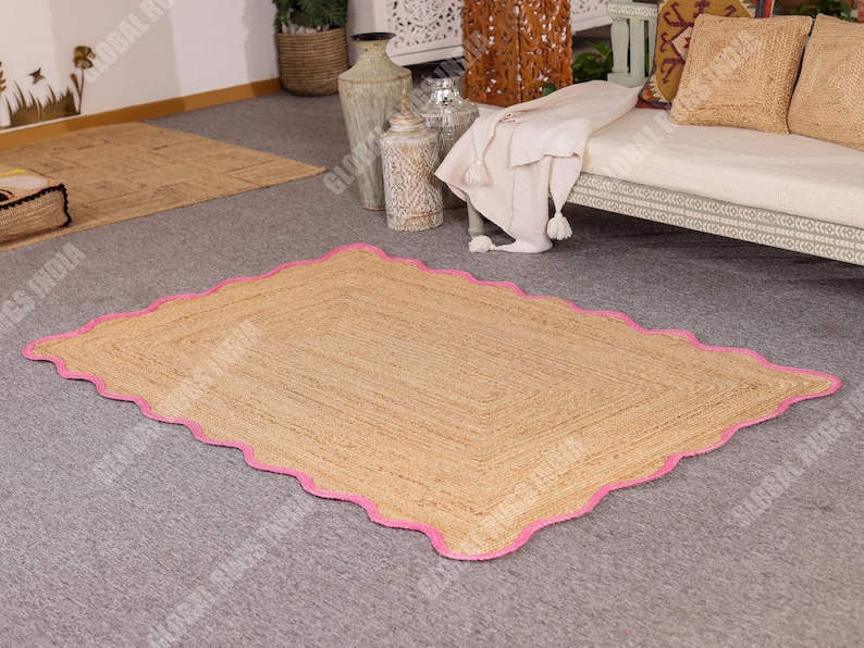 May include: A rectangular jute rug with a scalloped pink border. The rug has a natural tan color and is placed on a gray carpet. The rug's design features a woven texture, adding a rustic touch to the room's decor.