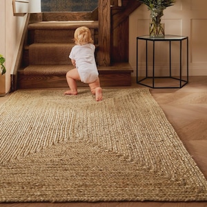 May include: A rectangular jute rug with a woven design, featuring a lighter border. The rug is placed on a wooden floor, with a baby in a white onesie crawling towards a staircase. The stairs are made of wood and have a brown carpet.