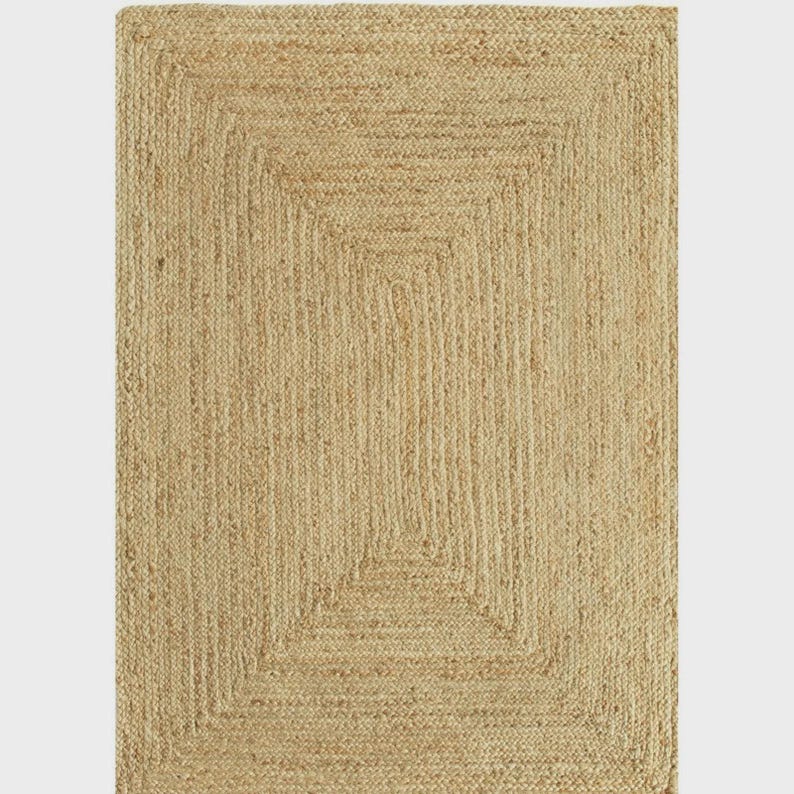 May include: Rectangular jute rug with a natural, light brown color. The rug features a woven design with a rectangular border and a central rectangular pattern. This rug is suitable for various home decor styles.