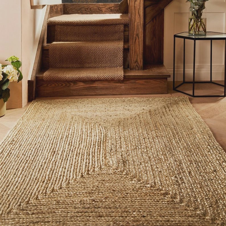 May include: A natural fiber rug with a geometric design lies on a wooden floor in front of a staircase. The rug is rectangular with a lighter-colored square border and a central design. The stairs have a brown carpet runner.