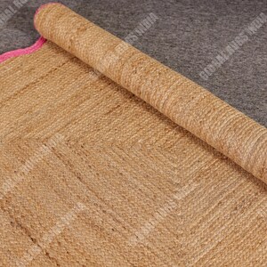 May include: A rolled-up, natural-toned jute rug with a pink scalloped border. The rug has a woven texture and a geometric pattern. The rolled portion reveals the rug's thickness and construction. The rug is on a gray surface.